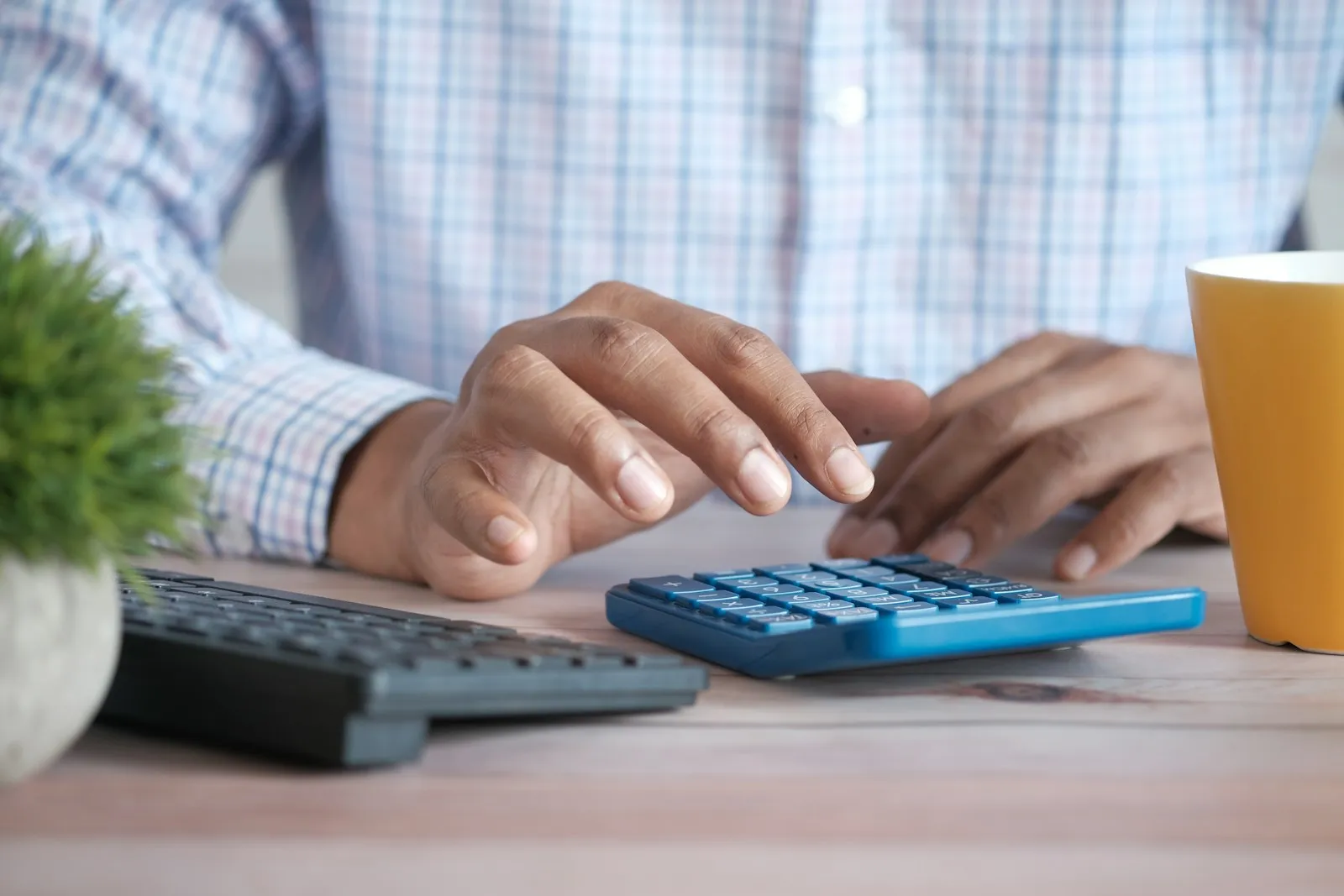 Close-up of hands tapping a blue calculator on a wooden desk beside a black keyboard, a yellow mug and a small potted plant