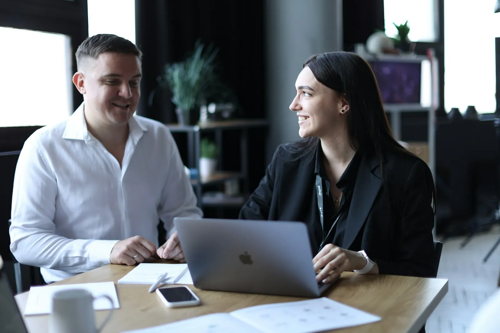A man and a woman in business attire smiling during a meeting at a table with a laptop in a modern office
