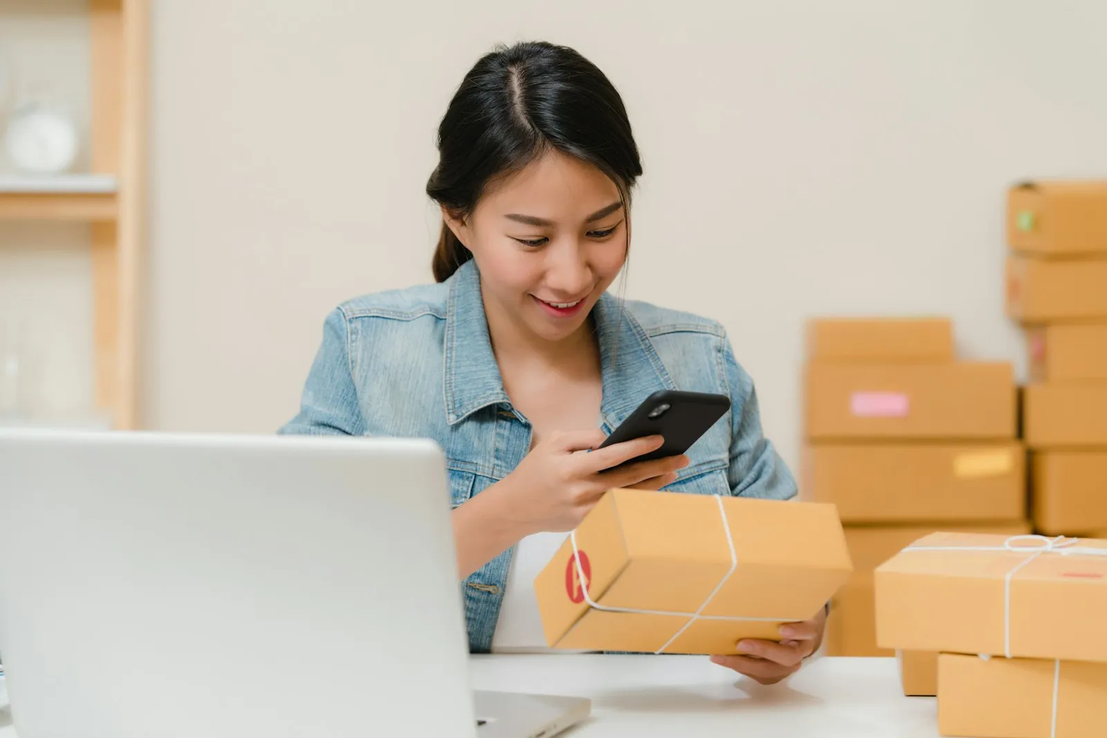 A smiling woman in a denim jacket checks her phone while holding a cardboard parcel, with a laptop on the desk and shipping boxes stacked behind her