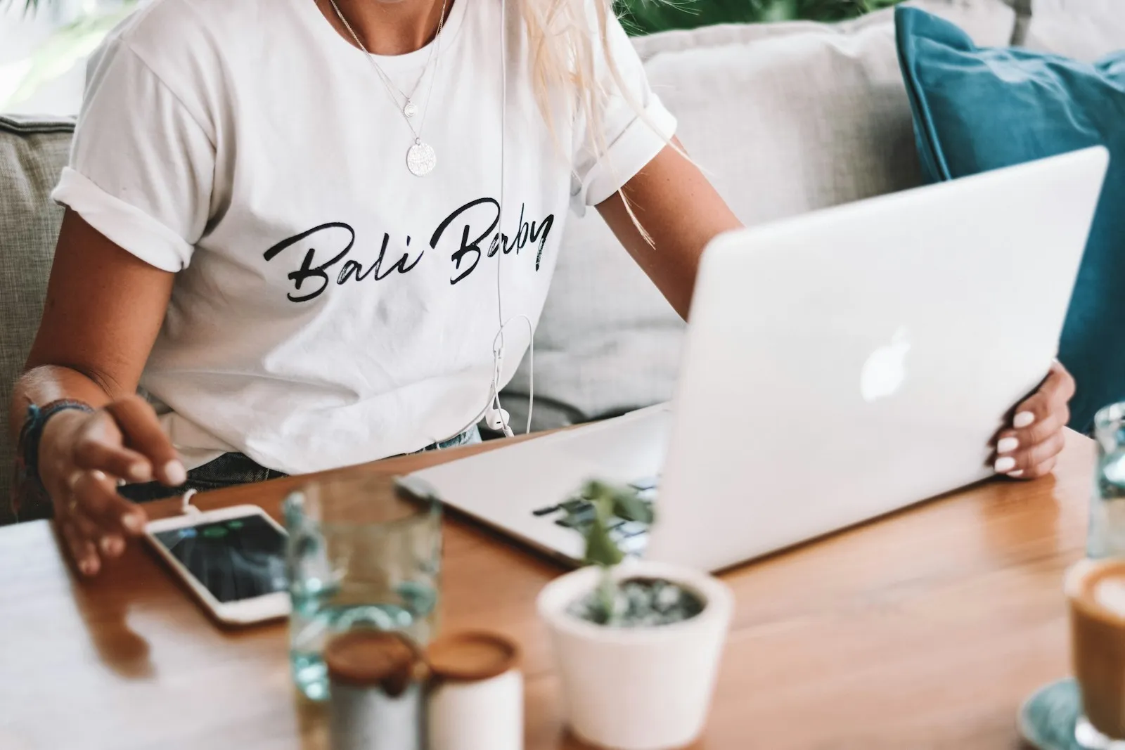 A woman in a white T-shirt working on a laptop at a wooden table, with a phone and a small potted plant beside her