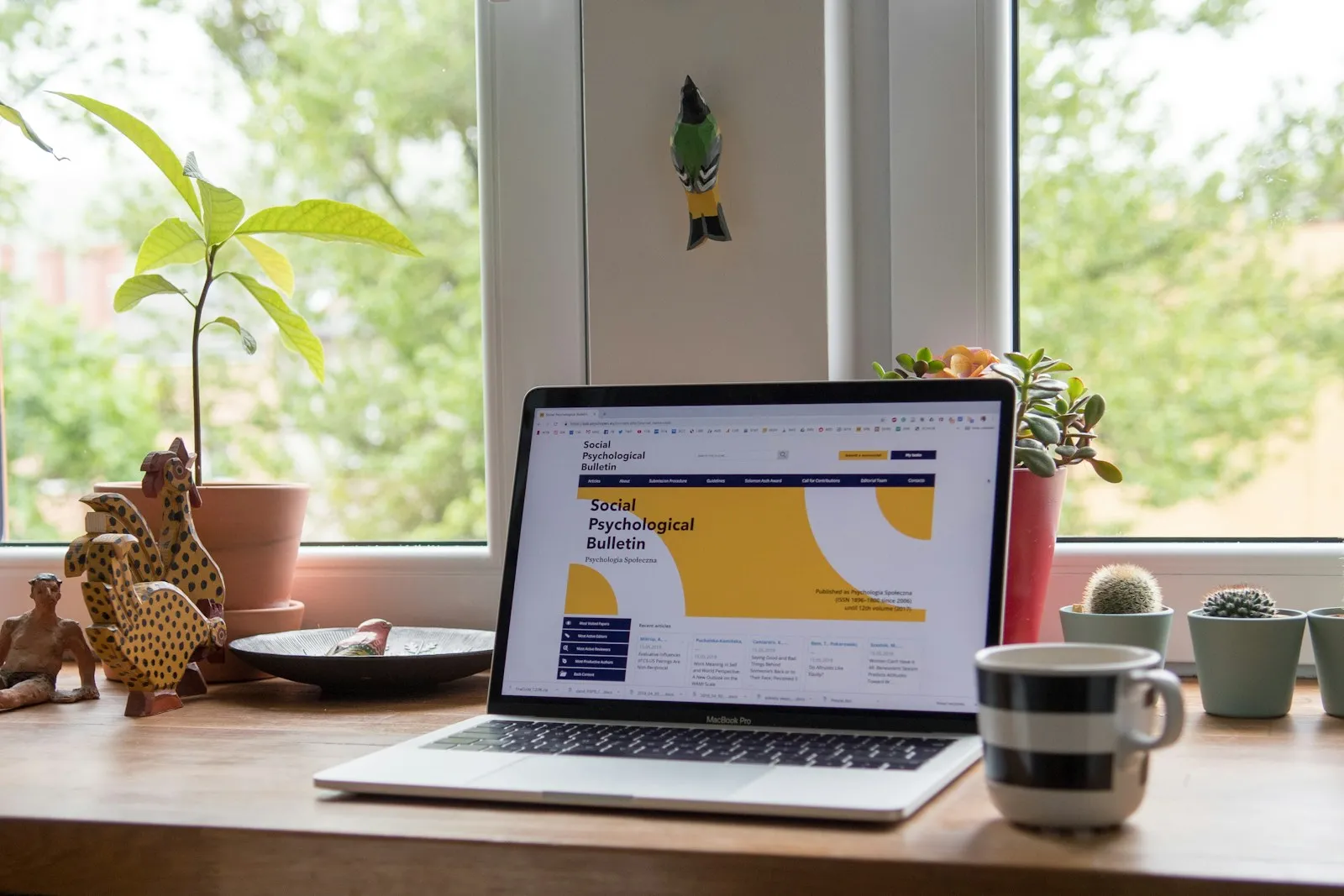 An open laptop showing a website on a wooden desk by a bright window, surrounded by potted plants, small cacti, carved wooden birds and a striped coffee mug