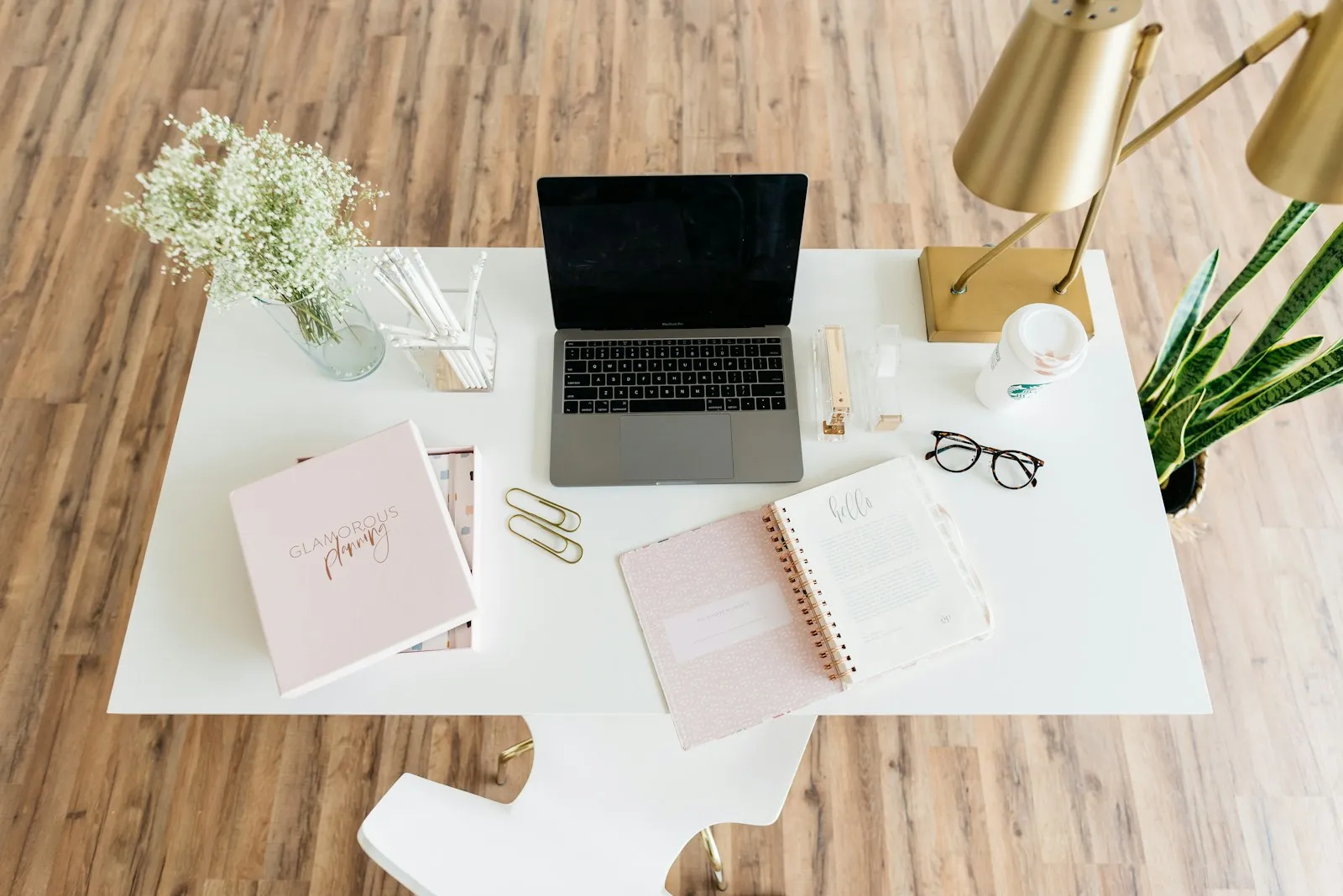 Minimalist white desk seen from above with an open laptop, a spiral planner, a cup of pencils, glasses and a small vase of white flowers