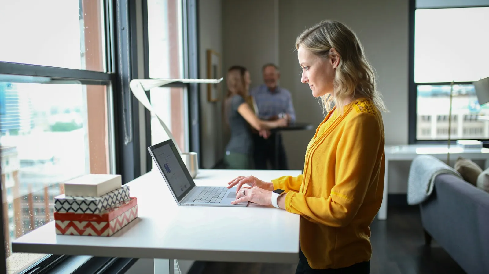 A woman in a yellow top typing on a laptop at a standing desk beside a bright office window