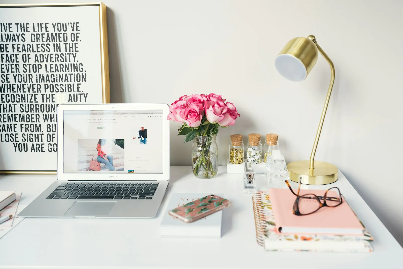 A MacBook Air beside a gold desk lamp, pink roses and spiral notebooks on a white desk